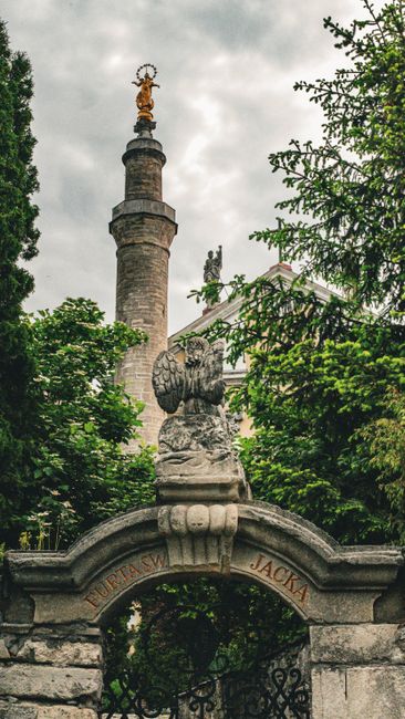 An outdoor scene featuring a stone archway topped with a decorative stone eagle figure, part of a garden entrance. Behind the arch, there is a well-maintained lawn with neatly trimmed grass and a vari